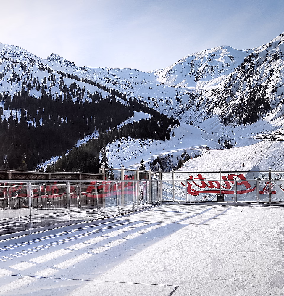 Terrasse mit Glasgeländer mit Blick auf einen schneebedeckten Berg mit Nadelbäumen.